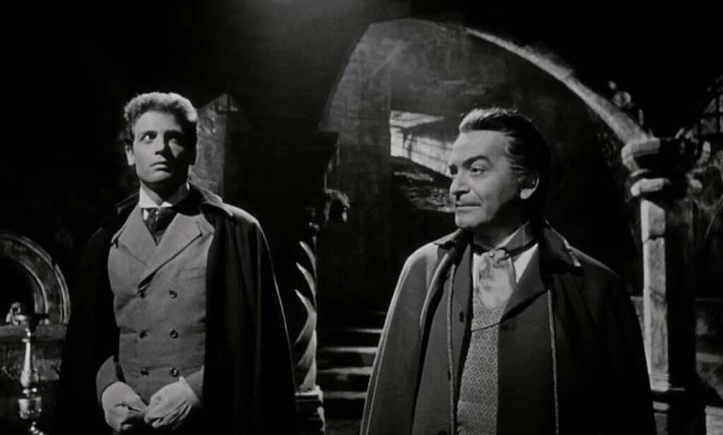A young man and an older man stand side by side in a spooky tomb. The young man looks apprehensive. The old man looks like he's about to cause trouble.