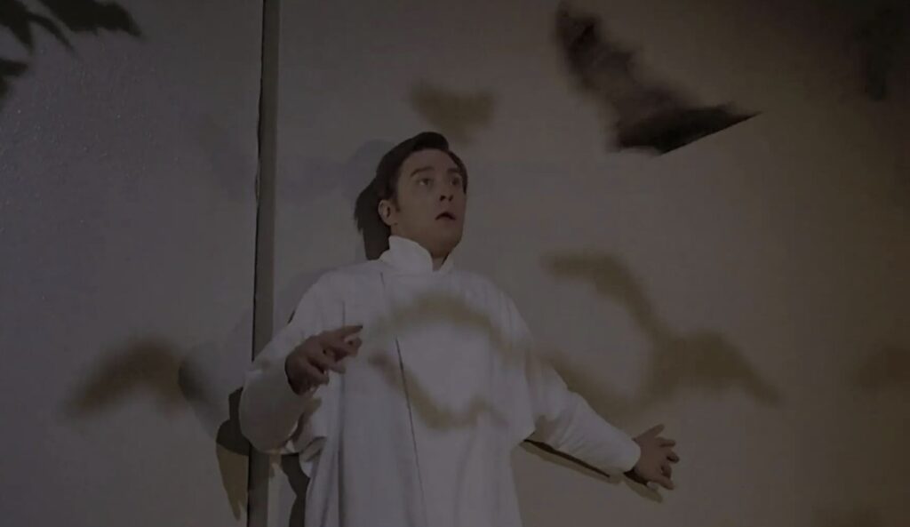 A young man in a white robe watches with a kind of stupified expression as a bat puppet swoops towards him. The shadows of more bat puppets are across him and the wall.